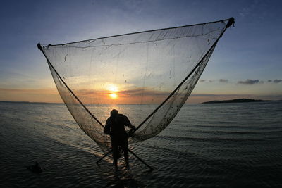 Silhouette fisherman with fishing net in sea against sky during sunset