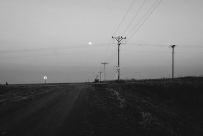 Dirt road by electricity pylons against sky at dusk