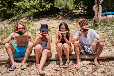 Company of friends sitting on tree log in forest and eating delicious watermelon while enjoying summer holiday together