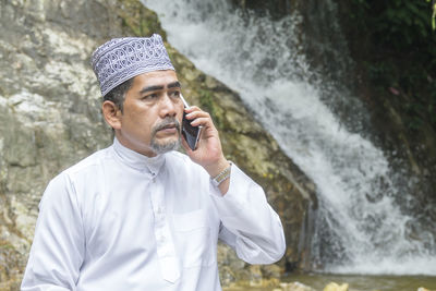 Thoughtful man talking on phone while sitting against waterfall