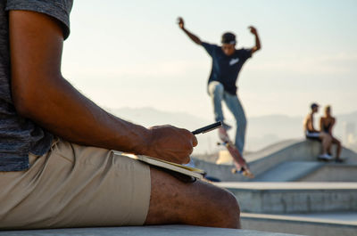 Midsection of man writing in book with friend skateboarding in background