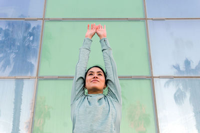 Confident young ethnic female athlete in stylish sportswear looking away while standing against modern glass building on city street