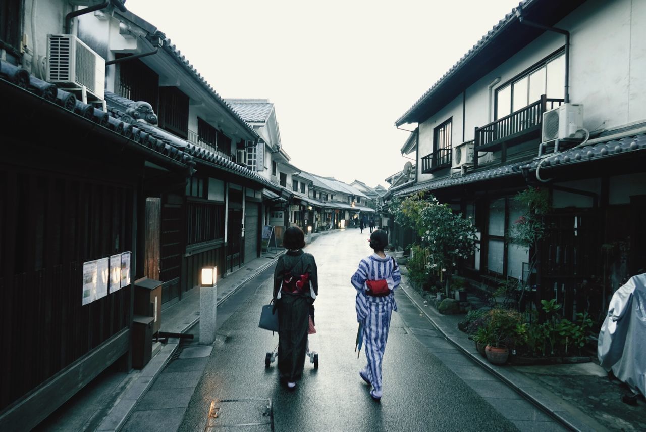 REAR VIEW OF PEOPLE WALKING ON ROAD AMIDST BUILDINGS IN CITY