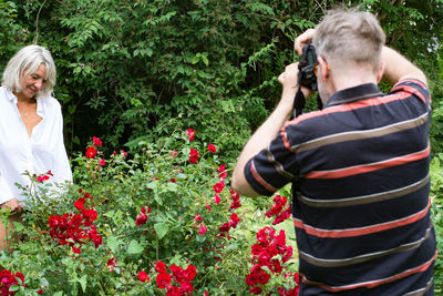 Rear view of man photographing woman standing by flowering plants