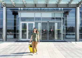 Young woman traveler carrying a yellow suitcase next to the entrance to the airport outside luggage