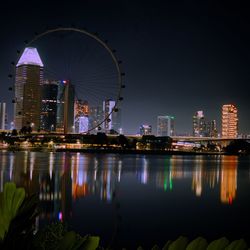 Illuminated ferris wheel by buildings against sky at night