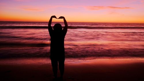 Silhouette woman forming heart shape at beach during sunset