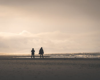Rear view of couple walking at beach against sky during sunset