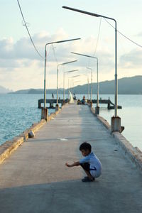 Boy crouching on pier over sea against sky