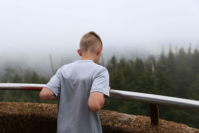 Rear view of teenage boy standing against forest during foggy weather