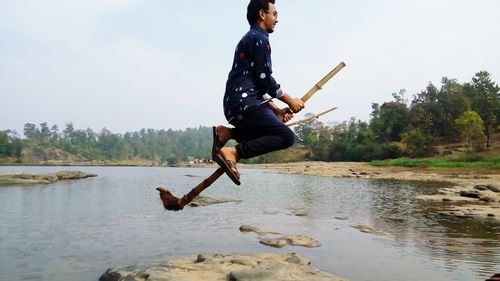 Side view of young man levitating with broom over lake against sky