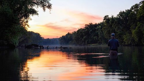 Rear view of man standing by lake against sky during sunset