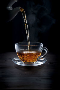 Close-up of teapot pouring drink in cup on table against black background