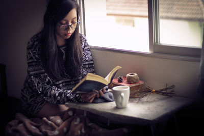 Woman reading book while sitting by window at home