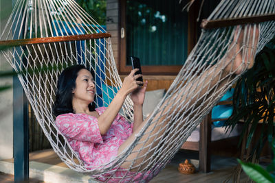 Happy asian woman making video call on smart phone while lying on hammock 