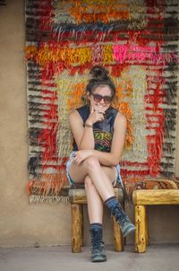 Portrait of smiling young woman wearing sunglasses while sitting on stool by wall