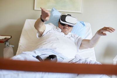 Surface level of senior man with arms raised using vr glasses on bed in hospital ward