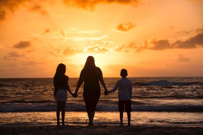 Rear view of mother with son and daughter standing at beach against sky during sunset