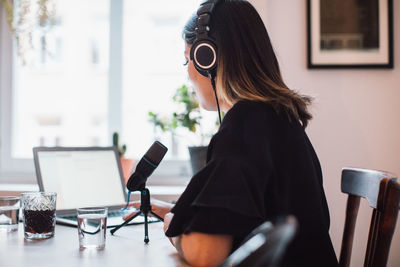 Side view of female podcaster wearing headphones sitting at table with microphone at home