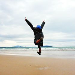 Rear view of cheerful man jumping at beach against cloudy sky