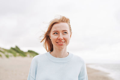 Portrait of young red haired woman in light blue sweater on sand beach by sea in storm