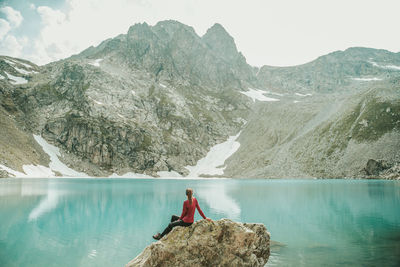 Man sitting on rock by lake against mountains