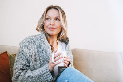 Young beautiful woman with blonde hair in cozy knitted grey sweater with cup of tea in hands at home