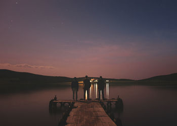 Rear view of men standing on pier by lake against sky at dusk