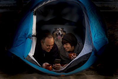 Couple with dog using mobile phone in tent at night