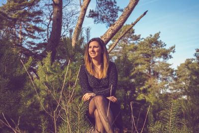 Young woman looking away while sitting on tree in forest