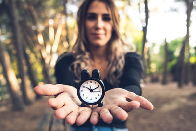 Portrait of young woman holding alarm clock against trees