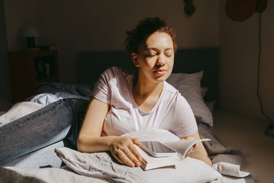 Teenage girl enjoying sunlight while while lying on bed with book at home