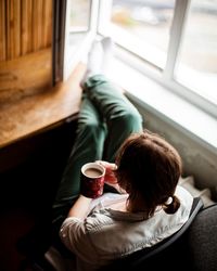 High angle view of woman having coffee while sitting by window at home