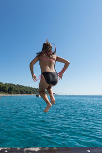 Rear view of young woman jumping in sea against clear blue sky