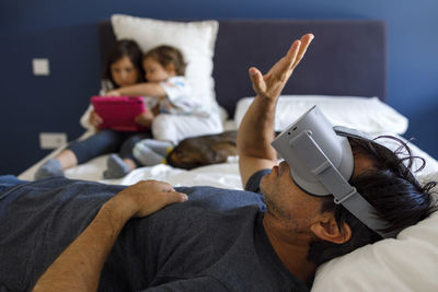 Man with virtual reality simulator gesturing while siblings using digital tablet on bed at home