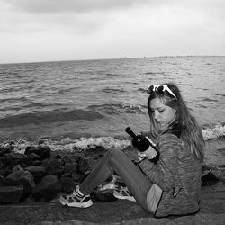 Side view of woman holding bottle while sitting at shore against sky