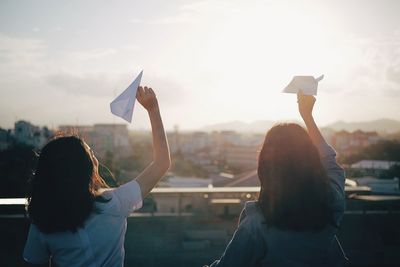 Rear view of women playing with paper airplanes against sky during sunset