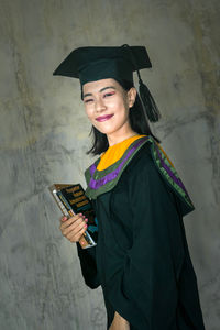 Portrait of smiling young woman holding smart phone while standing against wall