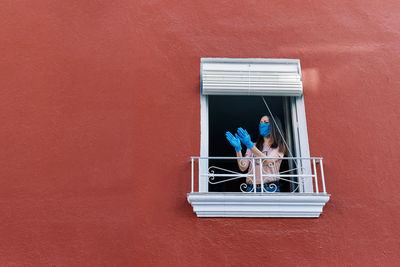 Young brunette woman with mask clapping from the window of the house with red background