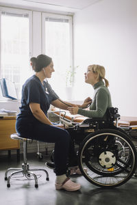 Female healthcare worker listening heartbeat of patient sitting on wheelchair in clinic