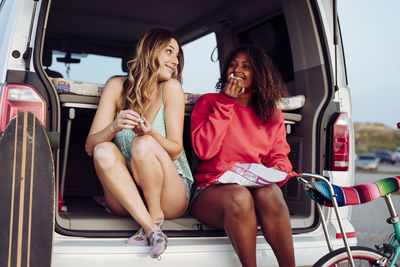 Cheerful women looking at each other while sitting on trunk of van eating popcorn