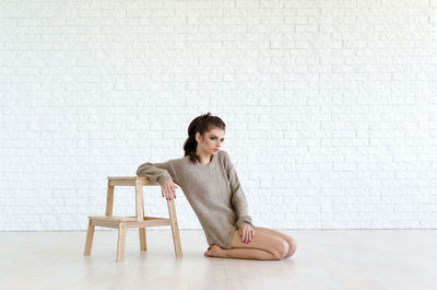 Full length portrait of young woman sitting by wood stool against brick wall