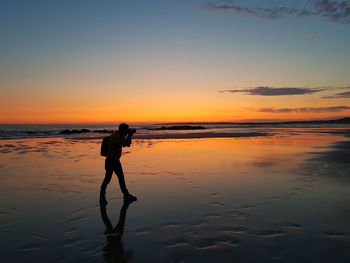 Silhouette man photographing while standing at beach against sky during sunset