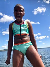 Cheerful teenage girl in bikini standing at beach against sky