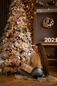 Cute teenage girl holding gift box sitting by christmas tree at home