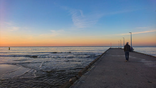 Rear view of man walking on pier over sea against sky during sunset