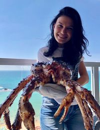 Portrait of smiling young woman holding large crab against sea and sky