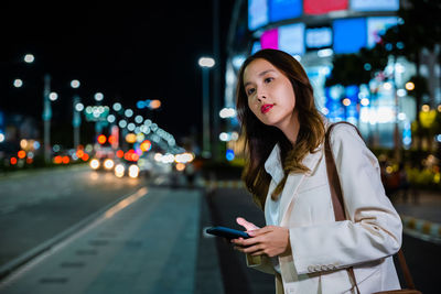 Portrait of young woman standing against illuminated city at night