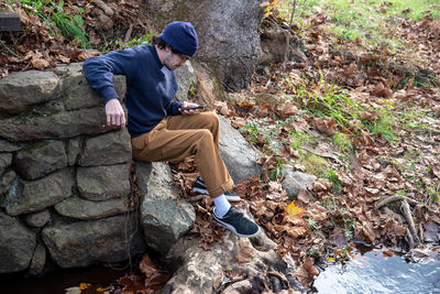 Young man checks phone in outdoor autumn nature scene by old stone wall and stream 