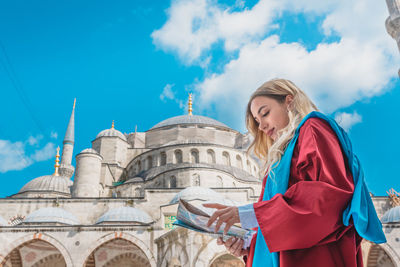 Low angle view of woman holding map outside mosque against blue sky